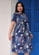 Woman with dark hair wearing a blue cotton dress with short sleeves and high round neckline in blue, white and red floral print, in front of blue door