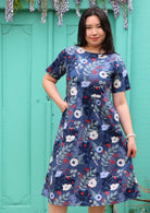 Woman with dark hair wearing a blue cotton dress with white and blue floral design, with hand in side pocket, in front of aqua door