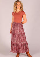 Model standing wearing 100 percent cotton three tiered maxi skirt in dark terracotta paired with scoop neck t-shirt and tan leather boots