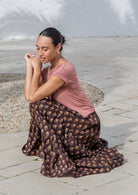 Model sits in tiered cotton maxi skirt in red, cream and light brown florals dark earthy base.