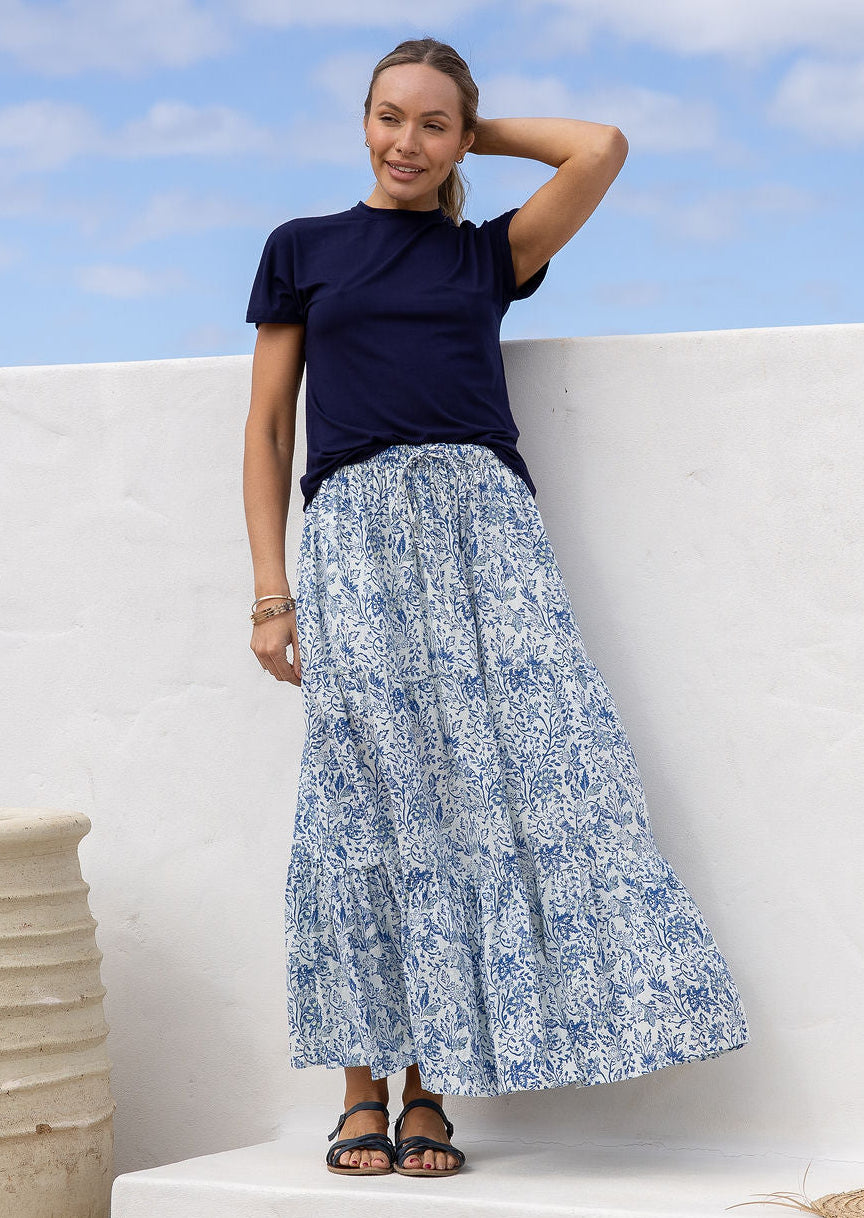 Woman wearing a white based blue floral cotton skirt standing against a white wall.