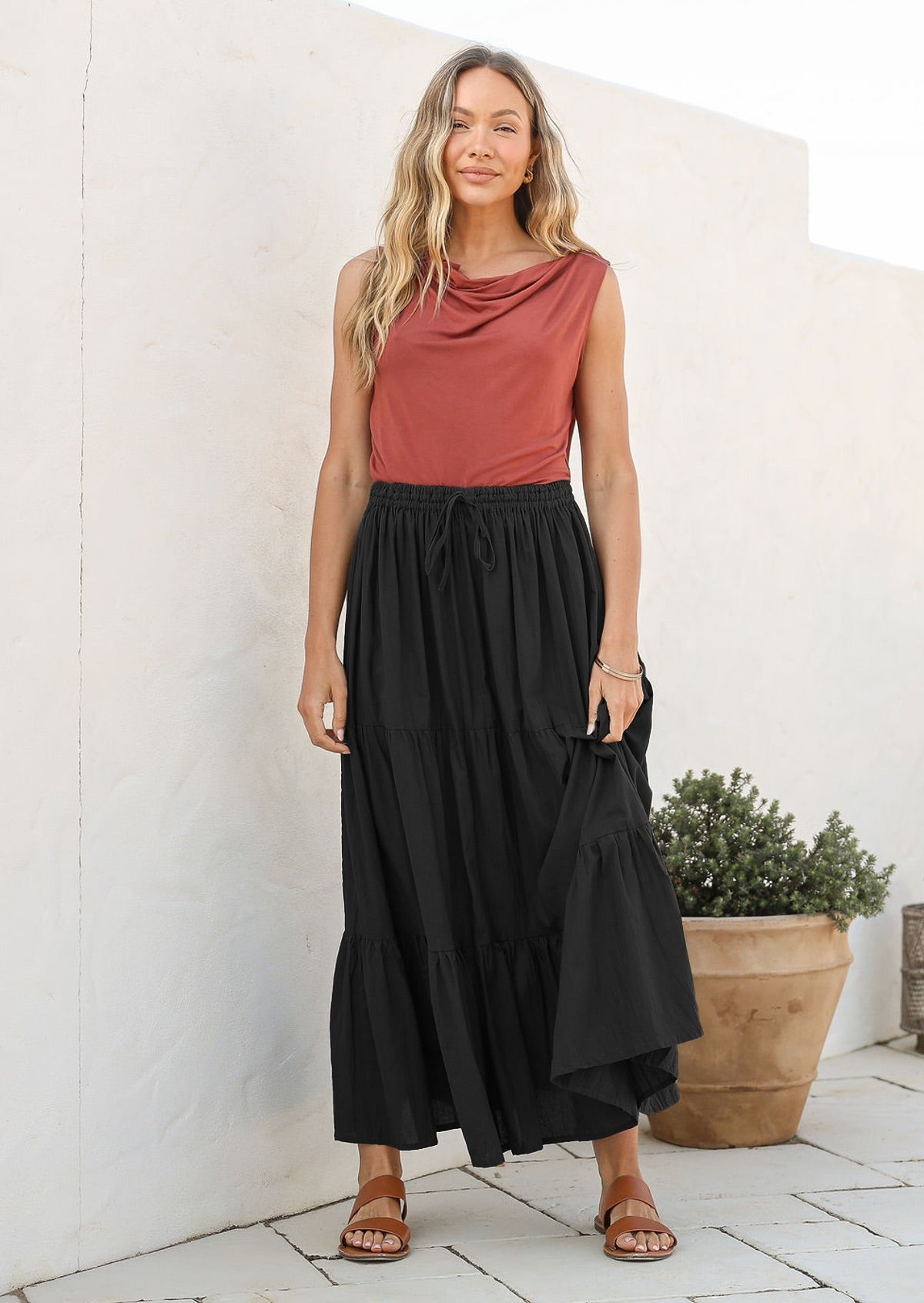 Woman wearing a dusty pink sleeveless top and black flowing cotton maxi skirt standing against a white wall.
