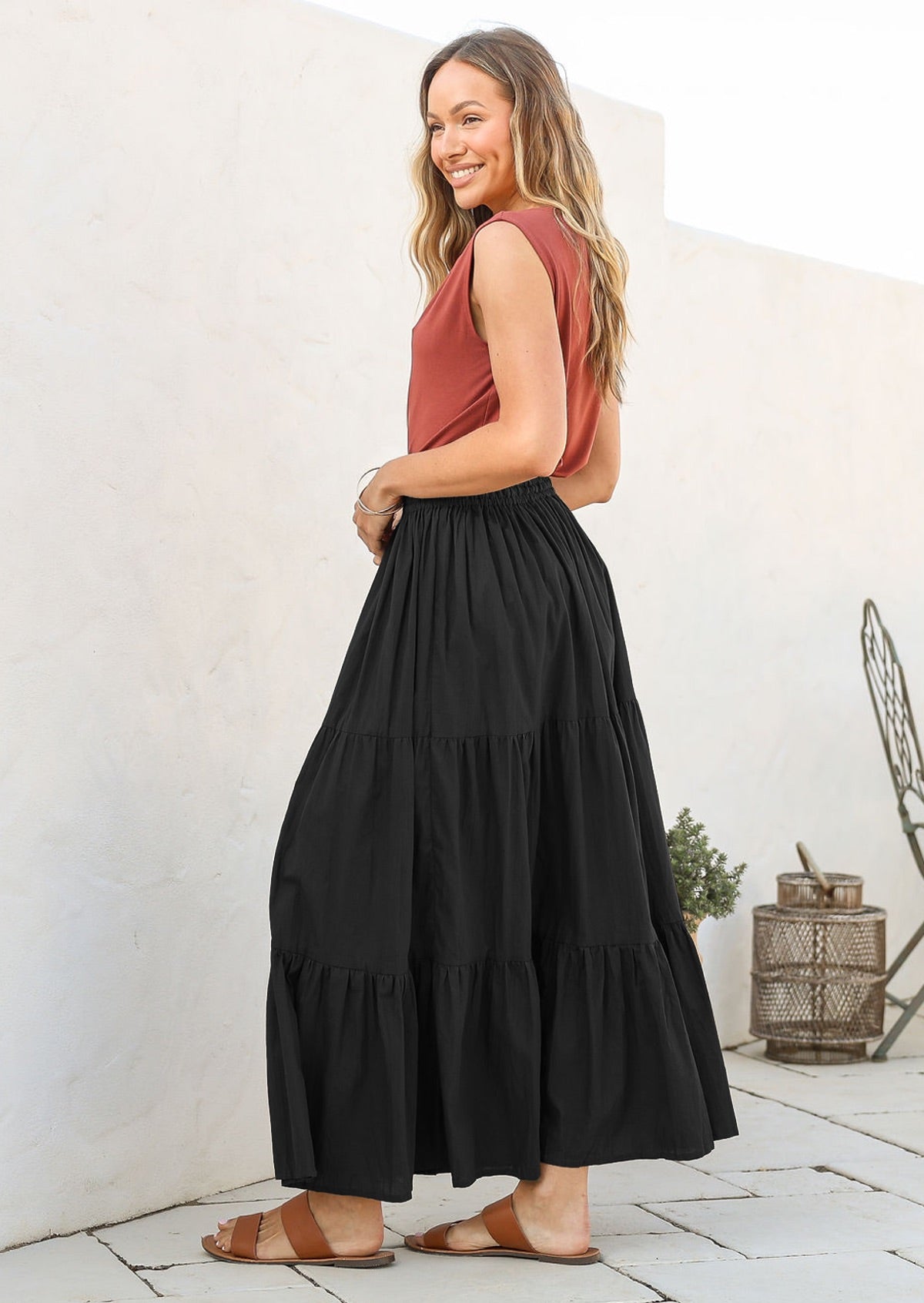 Woman standing side on wearing a dusty pink sleeveless top and black cotton tiered maxi skirt with tan sandals