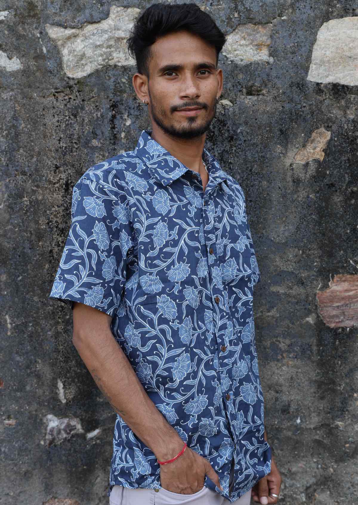 Man leans on wall in blue floral print cotton shirt with collar and coconut buttons