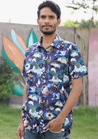 Man stands with hand in pocket, wearing a blue based floral print short sleeve shirt with birds interspersed