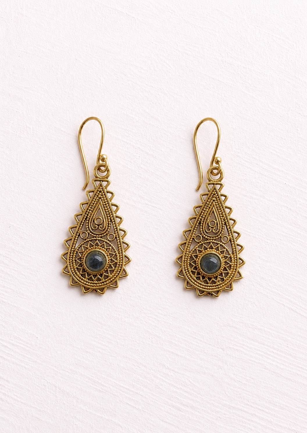 A pair of ornate gold-toned filigree dangle earrings with scalloped edges and a small round dark blue-gray stone at the centre of each earring, shown on a textured off-white background.