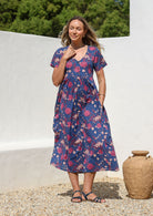 Woman standing outdoors with one hand in pocket wearing a relaxed fit, button through cotton dress in navy blue with V neckline and drawstring tie at the waist