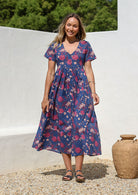 Woman standing in a 100% cotton floral print dress in blue with a V-neckline, short sleeves, pockets, and buttons through to the knee.
