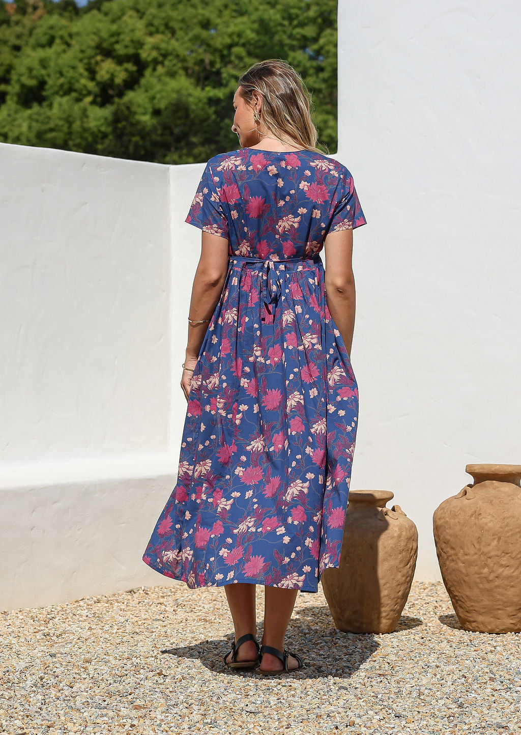 Back view of a model wearing a blue relaxed fit cotton dress with short sleeves, pockets, and a drawstring tie at the back.