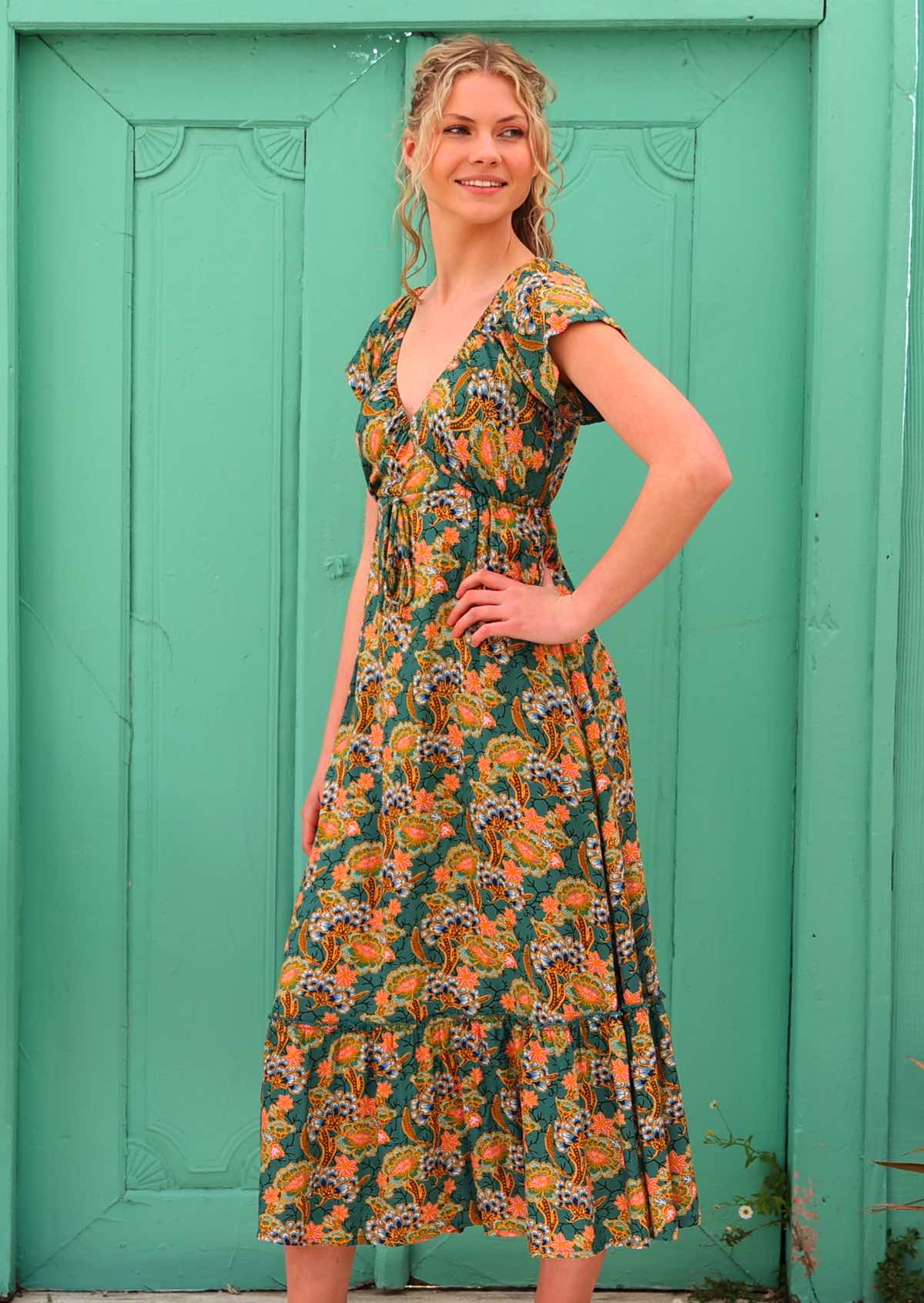 Model with hand on hip standing infront of aqua door wearing 100 percent rayon maxi dress in green based floral print