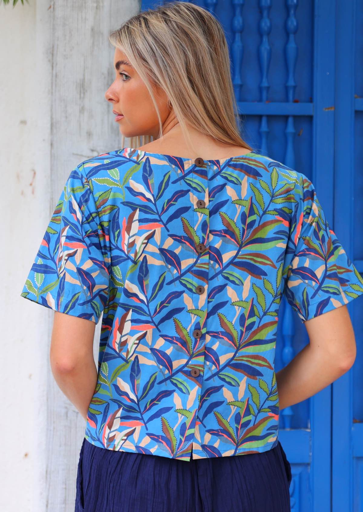 Woman wears blue 100 per cent cotton boxy blouse with multi coloured leaves with non functional decorative coconut shell buttons back view