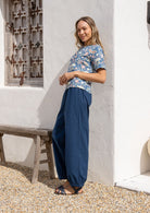 Model leans against a wall wearing a beautiful loose-fit, short-sleeved boxy cotton top in blue with delicate light pink, yellow, and green floral print 