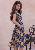 Woman wearing a 100% cotton, knee-length, A-line floral dress, with a beige wall and chair in the background.