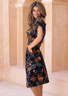 Model stands sideways with hands in pocket of a botanical print short sleeve sundress.