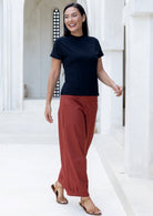 Woman wearing rust-colored cotton pants with pockets and pleated ankles, standing in a bright open area.