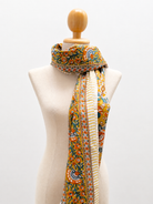 A multicolor patterned scarf with a cream center stripe and embroidered borders, draped on a beige fabric mannequin torso against a white background.