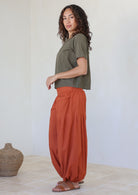 Woman wearing an olive green top and rust-colored pants standing against a white wall.