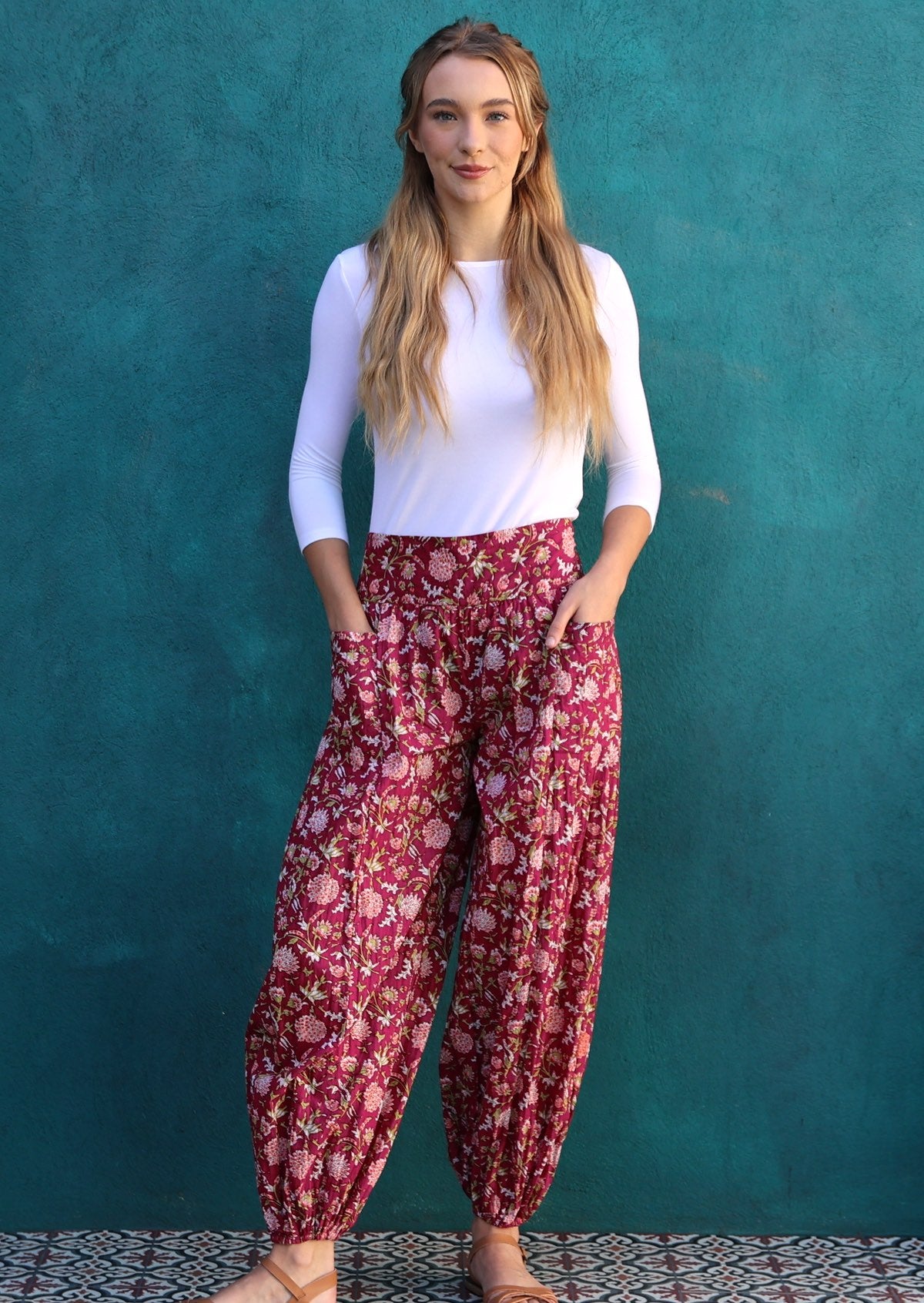 Model standing in front of blue wall wears harem style cotton pants in bright red colour with pink floral base.