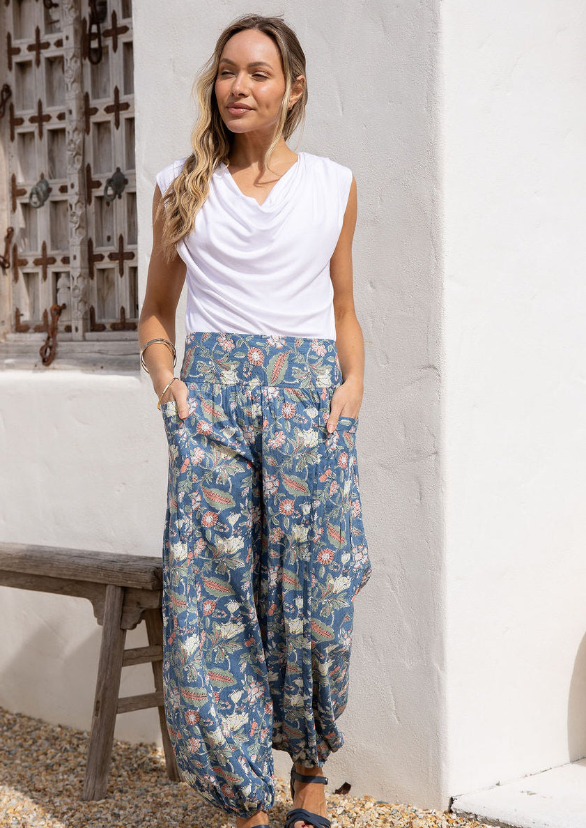 Woman wearing a white rayon top and a dusty blue floral pants with hands in deep pockets.