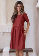 Woman poses in a burnt henna cotton dress with a round neckline, loose waist, above the knee length, and hands in pockets