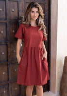 Woman wearing a rust toned, relaxed fit, above knee length cotton dress with short sleeves, pockets and round neckline.