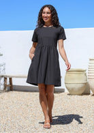 Woman wearing short sleeved black cotton with high round neckline, pockets and small gathers at the waist 