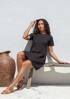 Woman in a above knee length black cotton dress sitting on a wooden bench against a white wall