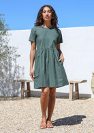 Woman with one hand in pocket wears 100% cotton above knee length loose fitting dress in jungle green shade