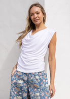 Woman wearing a white sleeveless top with pleats at each shoulder paired with floral pants against a light background