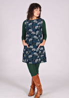 Woman stands in cotton corduroy sleeveless tunic layered over dark green leggings and long sleeve top. with hands in pockets