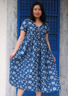 Model standing in front of blue door holding skirting of dress with both hands wearing 100 percent cotton navy blue button through v-neck dress with pockets