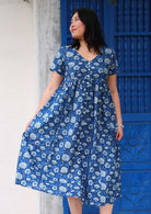 Model standing in front of blue door wearing 100 percent cotton navy blue button through v-neck dress with pockets