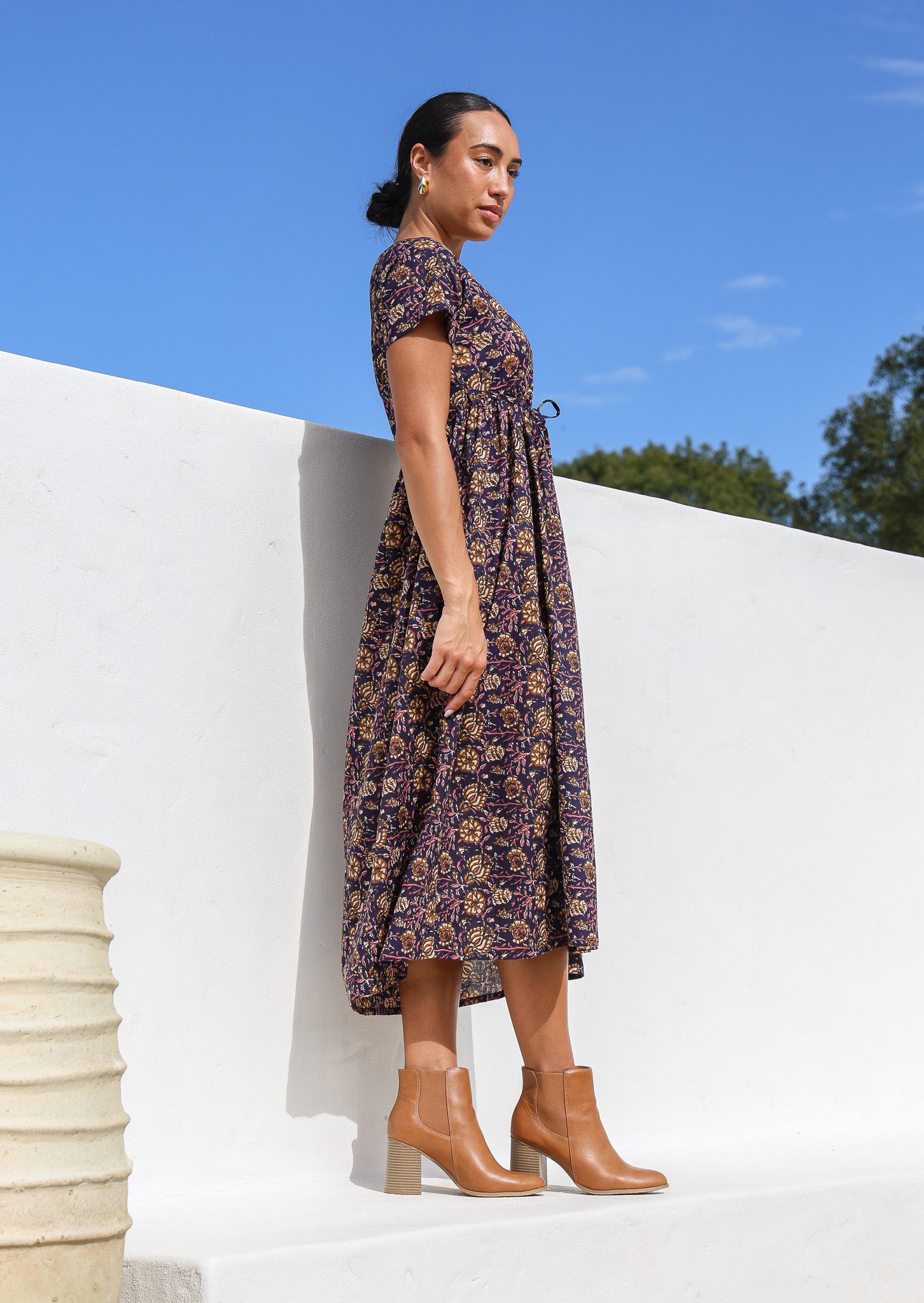 Model posing on side wearing a button through cotton dress with short sleeves and pink floral print on dark purple base.