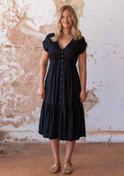 Woman stands in black rayon midi length dress with buttons through the centre front and elastic through the waistband