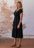 Woman standing sideways in black midi length rayon button through dress with V-neckline and elasticated waistband