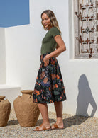 Model poses with hands in pockets wearing a knee length black cotton skirt featuring a lovely botanical print with belt loops, box pleats and elastic back waist