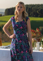 woman in retro inspired Scarlett Dress Dianthus in a  vibrant navy, purple, pink and aqua floral print on 100% cotton in front of laid out table 