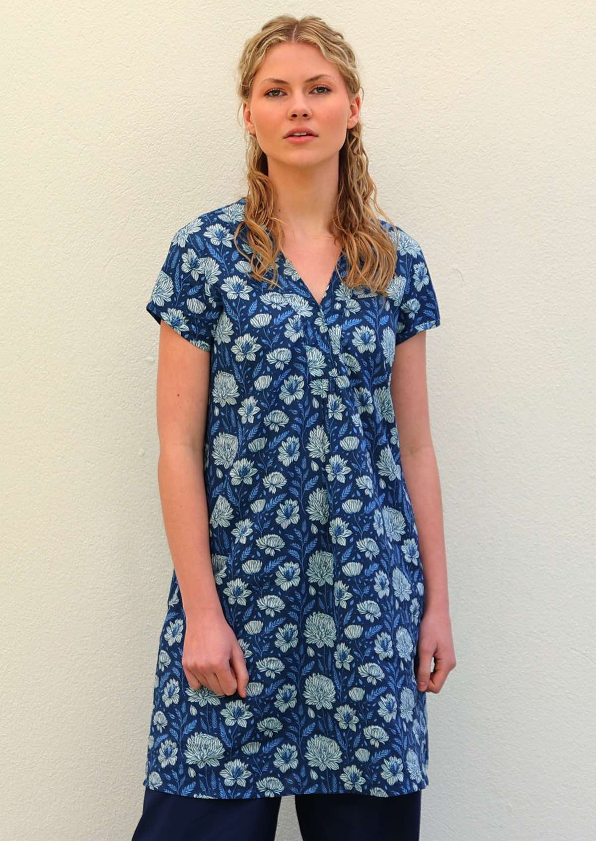 Woman with blonde hair wearing a blue cotton tunic dress with light blue and white floral design, in front of beige wall