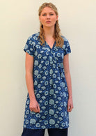 Woman with blonde hair wearing a blue cotton tunic dress with light blue and white floral design, in front of beige wall