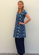 Woman with blonde hair wearing a blue cotton tunic dress in light blue and white floral print with blue cotton wide leg pants, in front of beige wall