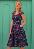 woman in retro inspired Scarlett Dress Dianthus in a  vibrant navy, purple, pink and aqua floral print on 100% cotton with hand in pocket in front of aqua door