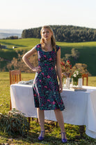 woman in retro inspired Scarlett Dress Dianthus in a  vibrant navy, purple, pink and aqua floral print on 100% cotton with hand in pocket in front of country outdoor table setting