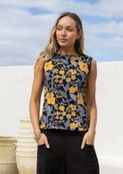 Woman standing in a retro floral sleeveless cotton top in black with plain trim around the arms.