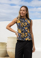 Woman wears a retro style sleeveless cotton top with yellow, pear green and grey florals on dark base 