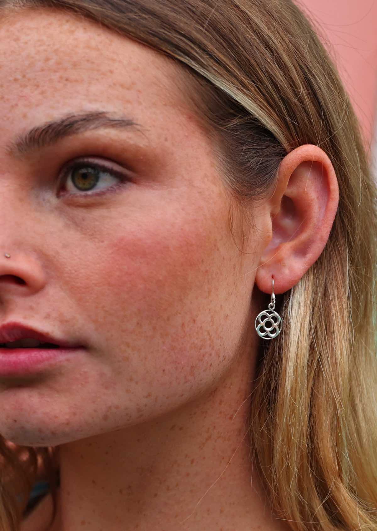 Woman wears silver disc earrings with four petal flower cutout, suspended from wire hook