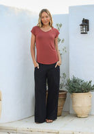 Woman wearing a 100% rayon terracotta V-neck top and black pants standing against a white wall with pot plants.