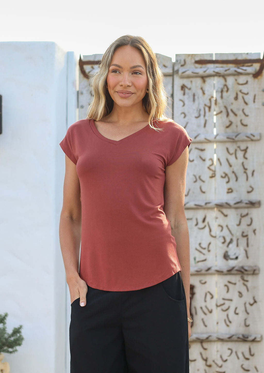 Woman with one hand in her pocket wearing a 100% rayon terracotta V-neck t-shirt and black pants standing against a textured wall.