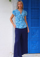 Full length photo of woman in blue floral cap sleeved top with hand in pocket of dark blue cotton wide legged pants