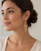 Model wearing long gold sunray drop earrings with an open geometric design, shown in a soft close-up portrait against a neutral background.