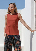 Woman standing with one hand in pocket wearing a 100% cotton sleeveless top in burnt henna with high round neckline and side bust darts